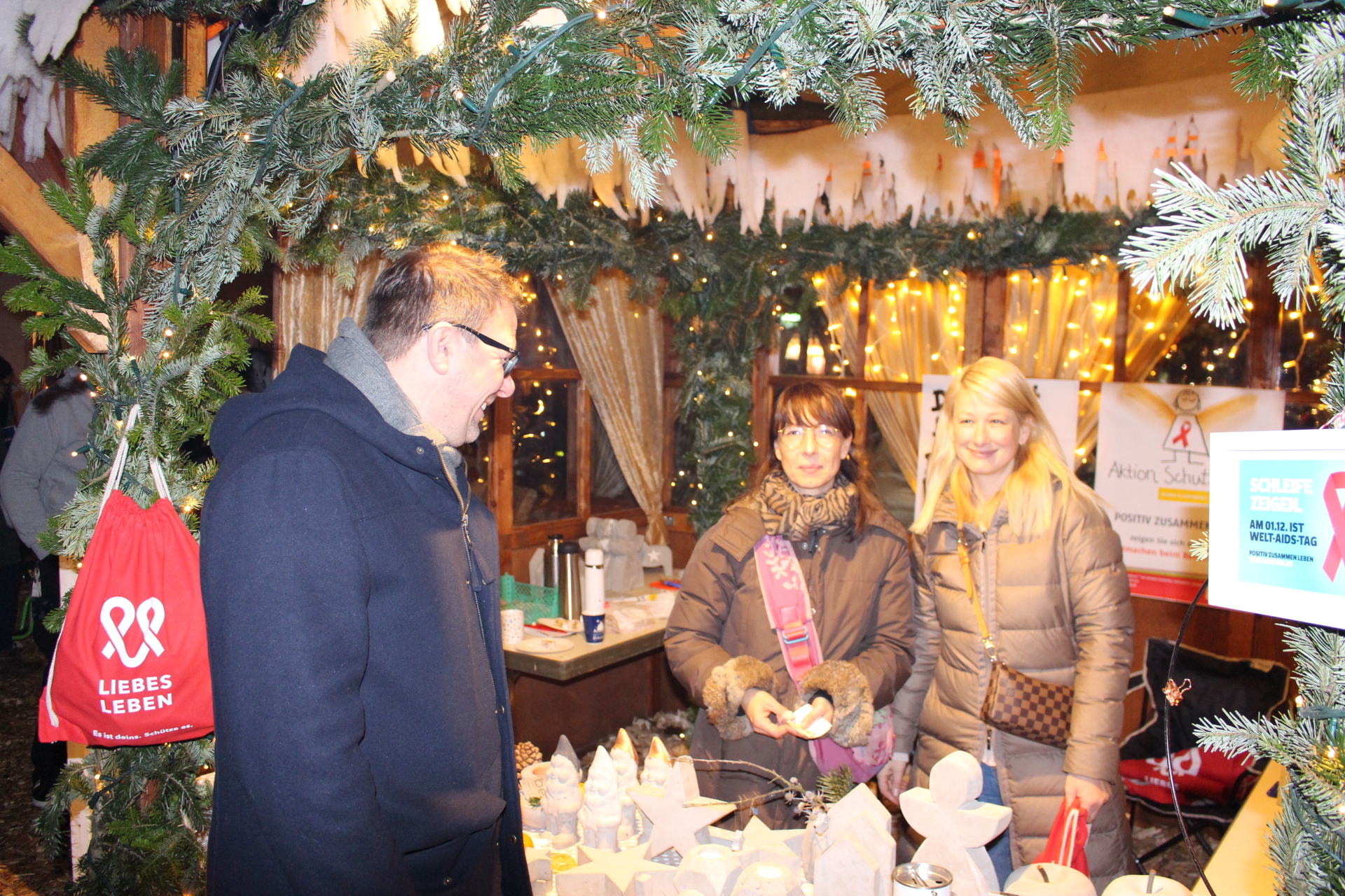 Ein Gast des Adventsdorfes steht an einem Verkaufsstand mit zwei Verkaufskräften