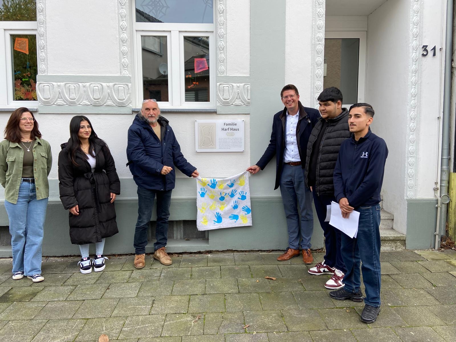 Kita-Leiterin Eva Jabs (l.), Hubert Rütten vom Heimatverein (3.v.l.) und Bürgermeister Stephan Muckel (3.v.r.) enthüllen gemeinsam mit Schüler*innen der Gemeinschaftshauptschule das neue Schild am "Familie Harf Haus".